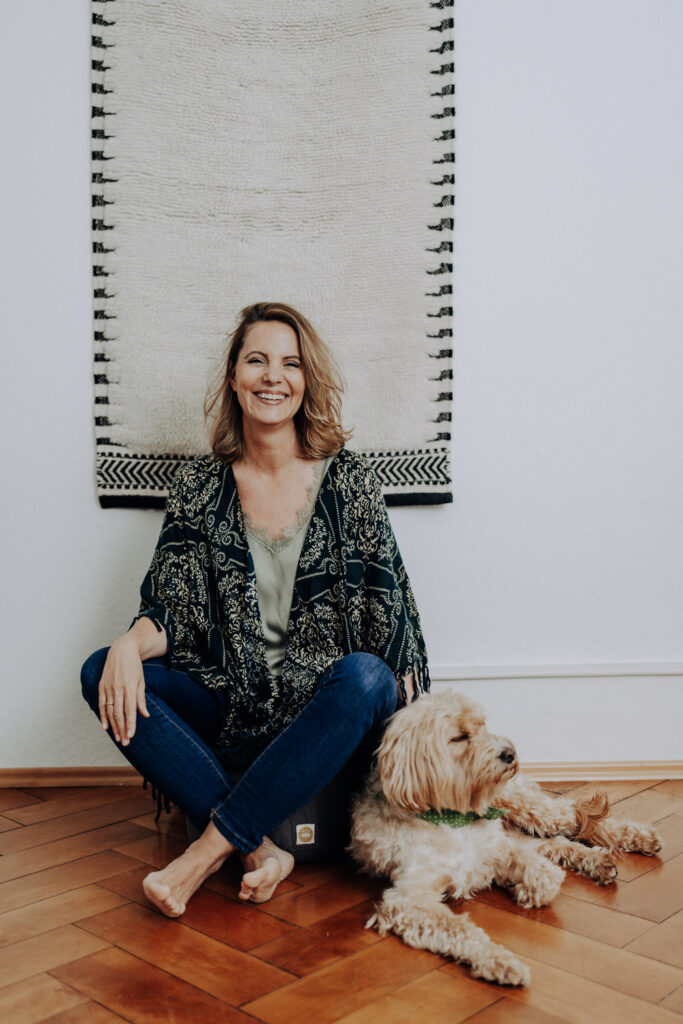 Personal Branding Foto einer Coachin, die vor einem Wandteppich auf dem Boden sitzt und ihre Hündin streichelt und in die Kamera lacht