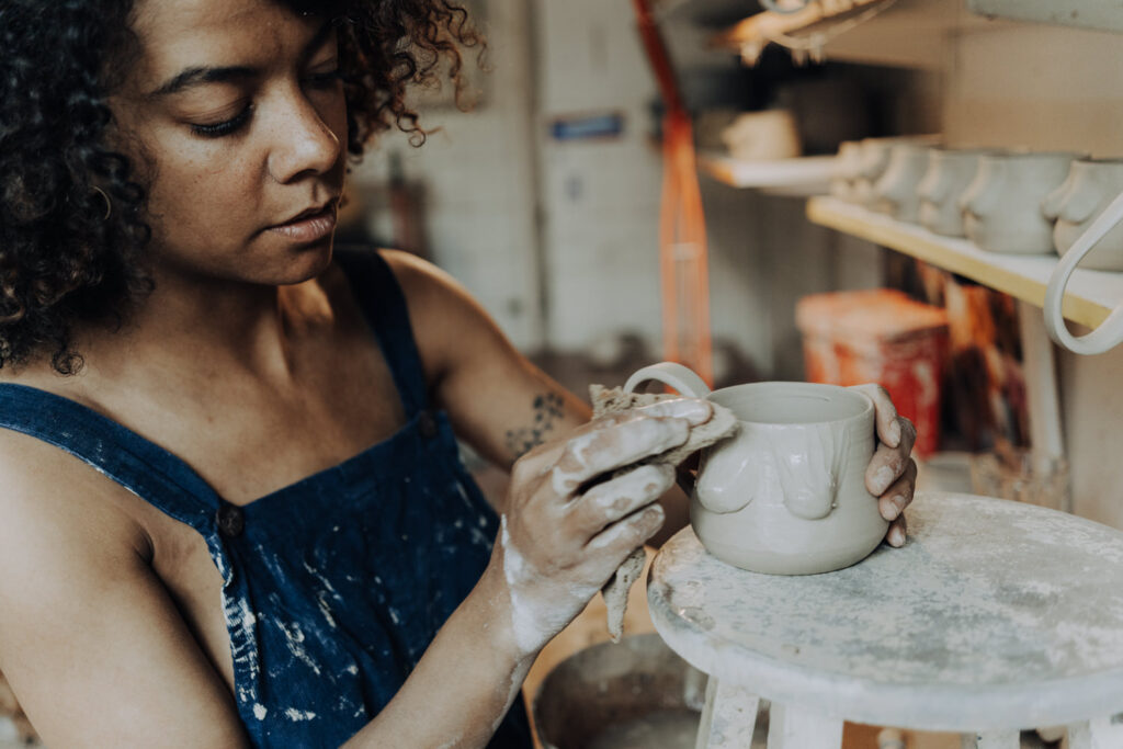 Businessfoto einer Keramikerin, die mit ihren Händen an einer Boobie-Tasse arbeitet. Personal Branding Foto einer Keramikerin, die eine unfertige Tasse mit einem Schwamm behandelt