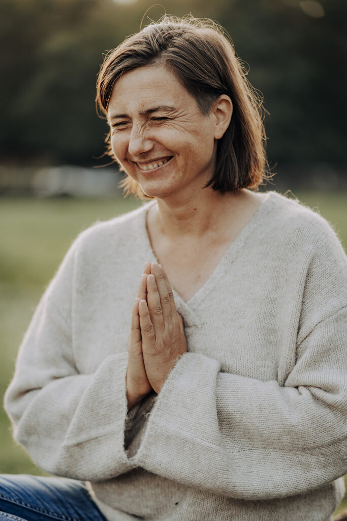 Frau zur Golden hour, grinst authentisch und hält ihre Hände in Gebetshaltung vor ihrem Körper Authentisches Businessfoto einer Hundetrainerin, die ihre Hände in der Gebetspose hat und mit geschlossenen Augen lacht